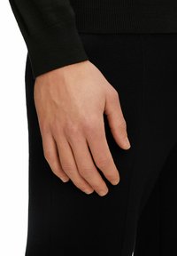 Schwarzer, gestrickter Pulloverärmel und eine Hand, die auf dunklen Stoffhosen ruht. Die Hand hat einen natürlichen Hautton und gut gepflegte Nägel.