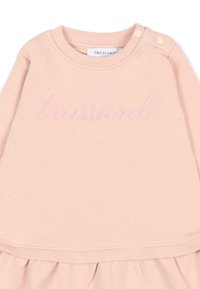Sweatshirt rose clair en tissu doux, à col rond et avec deux accents en boutons aux épaules. Présente un texte de logo dans une teinte de rose pâle.