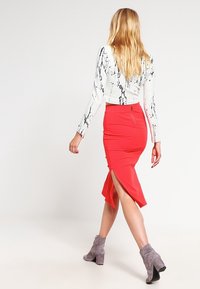 Roter, knielanger Bleistiftrock mit seitlichem Schlitz, kombiniert mit einem weißen Oberteil mit schwarzem Marmor-Muster. Graue Knöchelstiefel vervollständigen den Look.