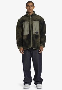 Chaqueta de forro polar con camuflaje verde y negro, dos bolsillos frontales, cierre de cremallera y tejido contrastante. Combinada con jeans oscuros y zapatillas blancas.