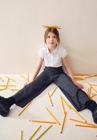 Chemise blanche à manches courtes et pantalon large gris foncé. Crayons jaunes et verts éparpillés sur un fond clair. Enfant assis avec des crayons équilibrés sur la tête.