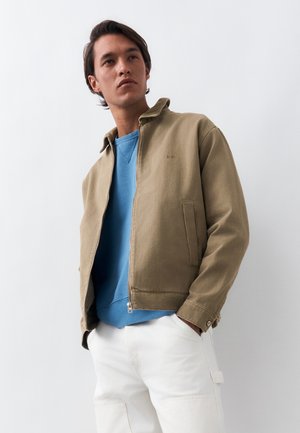 Młody mężczyzna w beżowej kurtce na zamek błyskawiczny, nałożonej na niebieską bluzę, i białych spodniach, stojący z rękami w kieszeniach na jednolitym tle.