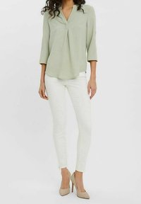 Femme portant un chemisier vert clair à manches trois-quarts, un pantalon blanc slim et des chaussures à talons beiges, debout devant un fond uni.