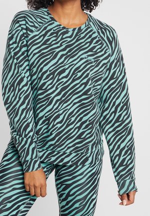 Mujer vestida con sudadera y leggings a juego con estampado de cebra en tonos teal y negro, de pie con una mano apoyada en el muslo.