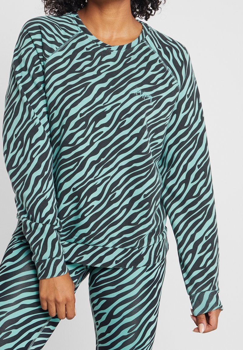 Femme portant un sweat-shirt et un legging assortis à motif zèbre teal et noir, debout avec une main reposant sur la cuisse.