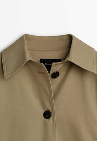Gros plan d’un col de manteau beige avec deux boutons noirs fermés, montrant une couture détaillée et une étiquette noire à l’intérieur de l’encolure.