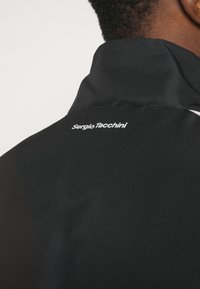 Melnas sporta jakas ar teksturētu audumu, ar augstu apkakli un baltu "Sergio Tacchini" logotipu aizmugurē. Minimālisma dizains.