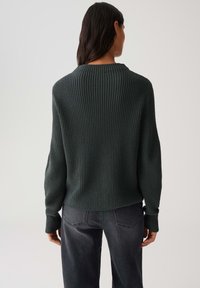 Donna con lunghi capelli scuri che indossa un maglione a coste verde scuro e jeans neri sbiaditi, in piedi con la schiena rivolta verso la telecamera su uno sfondo neutro.