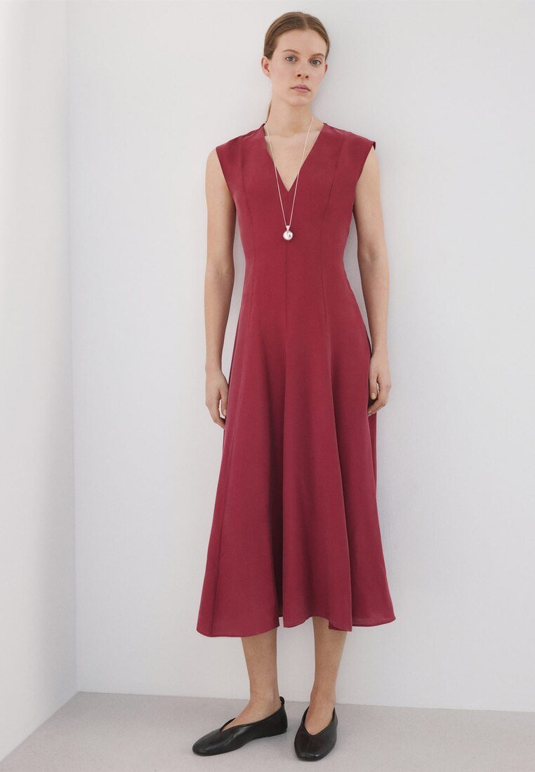 Femme portant une robe midi bordeaux sans manches et des ballerines noires, debout contre un mur blanc uni, avec un collier pendentif.