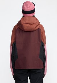 Veste à capuche bordeaux et marron avec des accents roses et noirs. Présente une texture lisse et imperméable ainsi qu'une coupe décontractée.