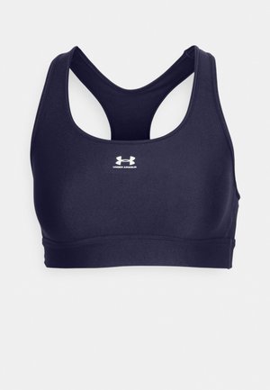 Tamsiai mėlyna sportinė liemenėlė su šoniniu dizainu, pagaminta iš glotnaus, tamprios medžiagos. Priekyje yra balta "Under Armour" logotipas.