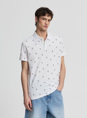 Giovane uomo che indossa una polo bianca con piccole stampe nere di palme e jeans azzurri, in piedi davanti a uno sfondo semplice.