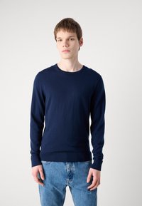 Pulover lung cu mânecă în culoare bleumarin, dintr-un material tricotat moale, având guler rotund și manșete și tiv cu nervuri, asortat cu blugi de culoare albastru deschis.