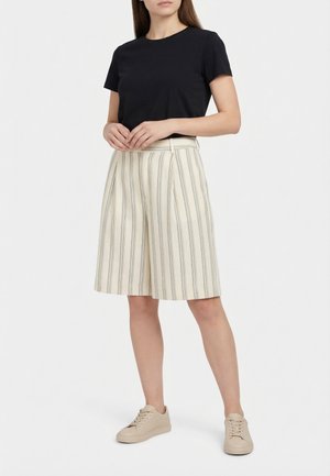 Mujer con una camiseta negra de manga corta, pantalones cortos crema de talle alto con rayas verticales y zapatillas beige, de pie frente a un fondo liso.