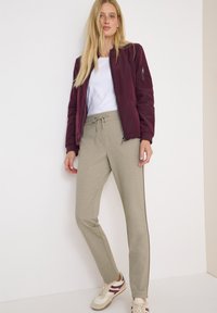 Frau steht an einer weißen Wand und trägt eine burgunderrote Bomberjacke, ein weißes T-Shirt, beige Jogginghosen mit Bindegürtel und beige Sneakers mit burgunderroten Akzenten.