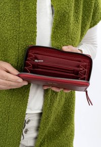 Manos sosteniendo una billetera abierta de cuero rojo con cremallera, que muestra múltiples compartimentos y ranuras para tarjetas en su interior.