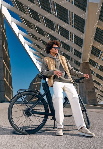 Elektrické kolo s matně černým rámem a silnými pneumatikami, zaparkované vedle osoby v šedé mikině, béžové vestě, bílých kalhotách a béžových botách.