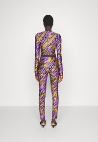 Lila tajt outfit med långa ärmar och byxor, prydd med gulddesign och svart text. Tillverkat av ett mjukt, elastiskt material.