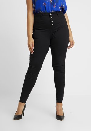 Frau trägt schwarze High-Waist-Skinny-Jeans mit Vier-Knopf-Verschluss, kombiniert mit schwarzen High Heels und einer blauen Bluse mit Blumenmuster.