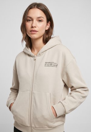 Junge Frau trägt einen beigen Kapuzenpullover mit Reißverschluss, Taschen und bedrucktem Text zur Selbstfürsorge auf der Brust, Hände in den Taschen, neutraler Hintergrund.