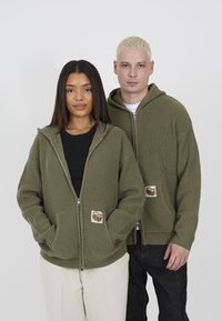Olive green zip-up hoodies in ribbed texture, featuring front pockets and a logo patch. Worn by two models against a white background.