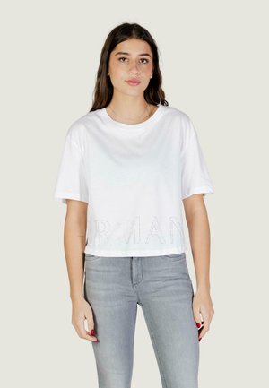 Jeune femme aux longs cheveux foncés portant un t-shirt blanc court et un jean gris clair sur un fond uni.
