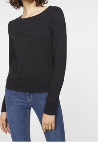 Kvinna iförd en svart långärmad topp med prickig textur och blå jeans, stående med vänster hand avslappnad vid sin sida.