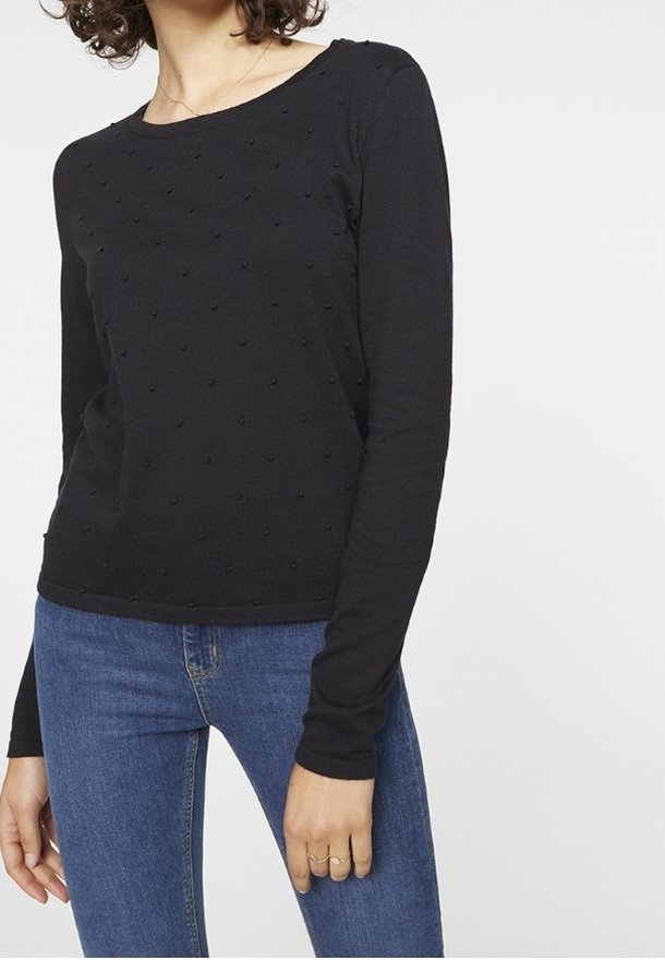 Kvinna iförd en svart långärmad topp med prickig textur och blå jeans, stående med vänster hand avslappnad vid sin sida.