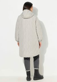Lys grå, quilted lang jakke med hætte og elastiske manchetter, lavet af syntetisk materiale. Båret over mørke jeans og sorte ankelstøvler.