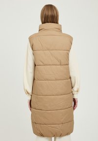 Lange, mouwloze donsbodywarmer in licht beige, voorzien van verticale stiknaden en een hoge kraag. De stof oogt glad en geïsoleerd.