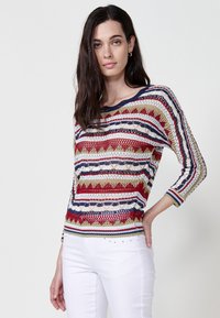 Pulover tricotat cu dungi și modele roșii, bleumarin, verzi și albe. Gât rotund, mâneci trei sferturi și o croială lejeră.