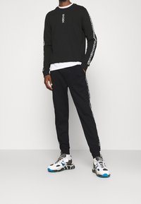 Černé tričko s dlouhým rukávem a bílým grafickým detailem na rukávech, doplněné černými tepláky s bočními pruhy a vícebarevnými teniskami.