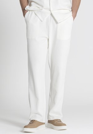 Persona con pantalones blancos acanalados y zapatillas beige con suelas blancas, de pie con las manos en los bolsillos frente a un fondo liso.