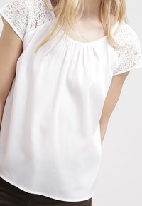 Blouse blanche avec manches courtes en dentelle, encolure ronde et tissu froncé au niveau de la poitrine. Matière lisse et légère avec une coupe décontractée.