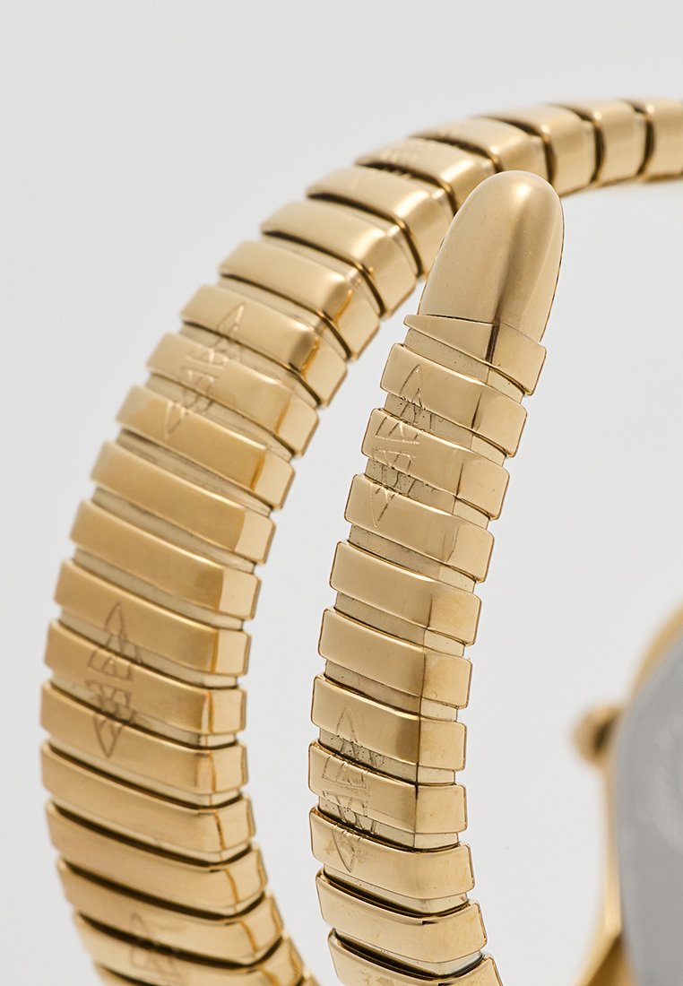 Montre en métal doré avec un bracelet en maillons texturé et flexible, et une extrémité lisse et arrondie. Présente des détails gravés sur les maillons. Cadran de la montre circulaire partiellement visible.
