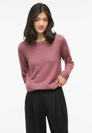 Mladá žena s tmavými vlasmi po plecia, oblečená v purpurovom svetri a čiernych širokých nohaviciach na bielom pozadí.