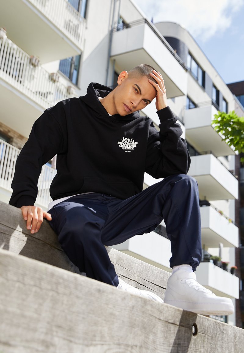Svart hettegensere med hvit grafisk tekst, parret med marineblå bukser og hvite sneakers, sitter på en treplattform utendørs.
