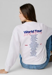 Νεαρή γυναίκα κάθεται φορώντας λευκό φούτερ με καταγραφή των πόλεων της Παγκόσμιας Περιήγησης Red Bull Racing 2026 στην πλάτη με μπλε και κόκκινο κείμενο.