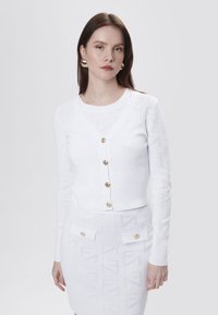 Witte gebreide cardigan met V-hals, vijf gouden knopen en een gestructureerd patroon. Gekozen in combinatie met een bijpassende witte rok met zakken en gouden accenten.
