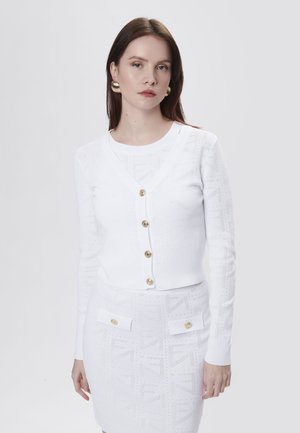 Witte gebreide cardigan met V-hals, vijf gouden knopen en een gestructureerd patroon. Gekozen in combinatie met een bijpassende witte rok met zakken en gouden accenten.