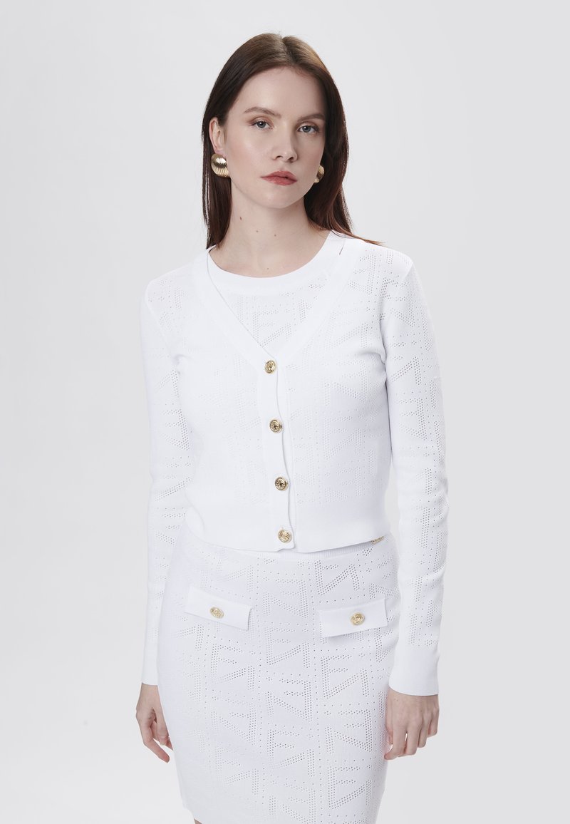 Witte gebreide cardigan met V-hals, vijf gouden knopen en een gestructureerd patroon. Gekozen in combinatie met een bijpassende witte rok met zakken en gouden accenten.