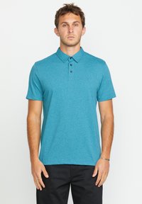 Türkises Poloshirt mit kurzen Ärmeln, drei schwarzen Knöpfen am Kragen und einem dezenten Texturmuster. Getragen mit schwarzen Hosen.