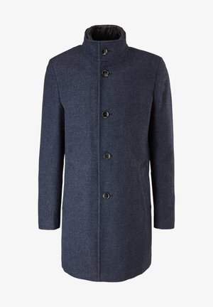 Mann in einem dunkelblauen Wollmantel mit schwarzen Knöpfen über einer schwarzen Zip-Jacke, stehend mit entspannten Händen an den Seiten.