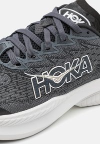 Baskets de sport en tissu gris avec un dessus en maille texturée, œillets renforcés et une marque blanche proéminente sur le côté. Semelle intermédiaire blanche incurvée.