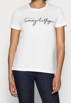 Kvinde iført en hvid T-shirt med korte ærmer med "Tommy Hilfiger" script-logo i sort, kombineret med mørkeblå jeans.