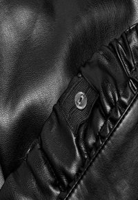 Černá koženka má strukturovaný povrch s nabranou částí a průhledným knoflíkovým detailem. Design obsahuje hladké i zmačkané oblasti.