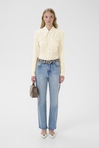 Langermet, lys gul bluse med rynket tekstur og knapper, kombinert med rette, lyseblå jeans og hvite spisse hæler. Holder en liten brun veske.