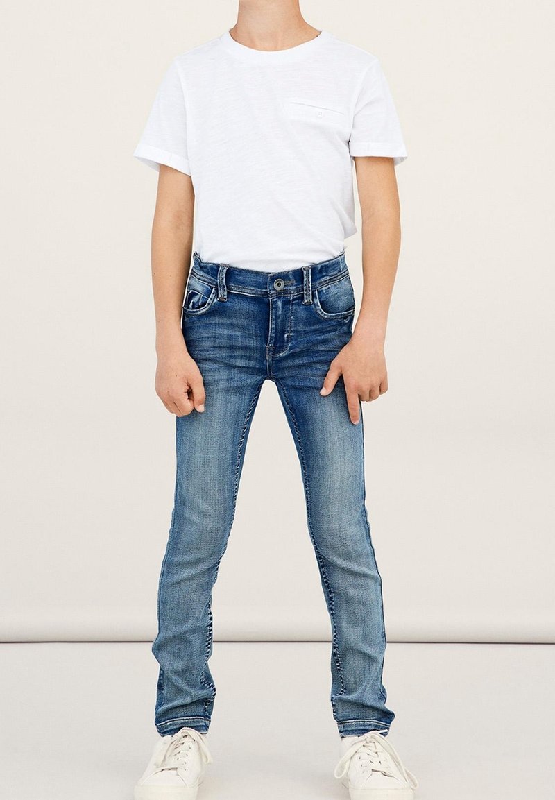 Enfant portant un t-shirt blanc uni à manches courtes, un jean slim bleu et des baskets blanches, debout devant un fond neutre.