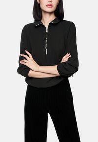 Zwart bovenstuk met ritssluiting en een hoge kraag, voorzien van raglanmouwen en elastische manchetten. Decoratieve logodetail aan de voorkant, gecombineerd met zwarte pantalon met krijtstrepen.