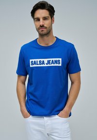 Camiseta corta de manga azul hecha de algodón, con un prominente logo blanco de "SALSA JEANS" en el frente. El modelo lleva pantalones blancos y tiene las manos en los bolsillos.